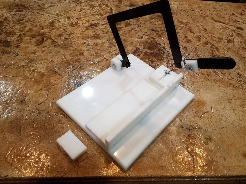 May include: A white plastic cheese cutting tool with a black metal frame and a wire for cutting. The tool is on a brown countertop.