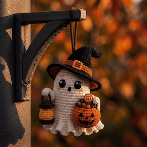 May include: A crocheted ghost ornament wearing a black witch hat with an orange band and buckle. The ghost has large black eyes, holds a small brown lantern in one hand, and carries an orange jack-o'-lantern bucket filled with candy.