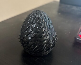 May include: A black, egg-shaped object featuring a textured, overlapping scale pattern across its entire surface. The structure is rounded at the base and tapers towards a point at the top, with visible ridges creating a layered, three-dimensional effect.