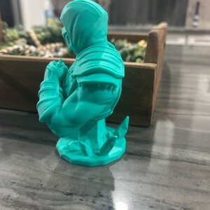May include: A bright turquoise 3D printed bust of a hooded warrior figure. The figure is shown from a side profile, displaying detailed shoulder armour, muscular arms, and a mask covering the lower face. The base features jagged, crystal-like elements.