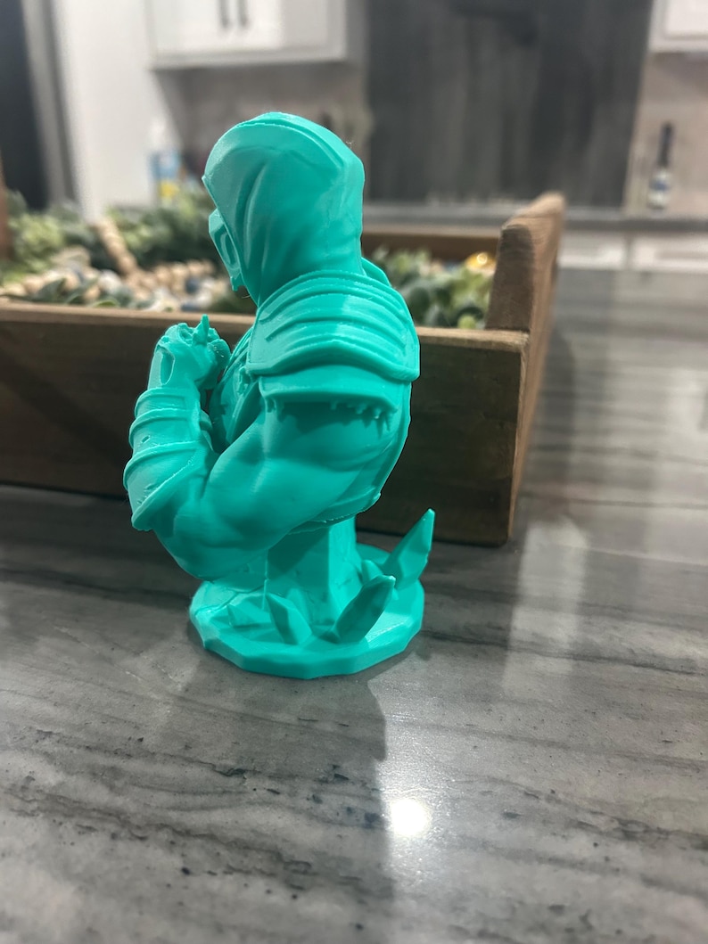 May include: A bright turquoise 3D printed bust of a hooded warrior figure. The figure is shown from a side profile, displaying detailed shoulder armour, muscular arms, and a mask covering the lower face. The base features jagged, crystal-like elements.