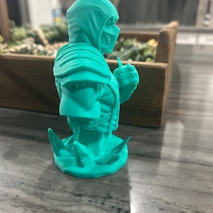 May include: A bright teal 3D printed bust of a hooded warrior figure wearing a mask and shoulder armour. The figure is depicted from the chest up, posed with a clenched fist, and stands on a base featuring jagged, crystalline-like protrusions.