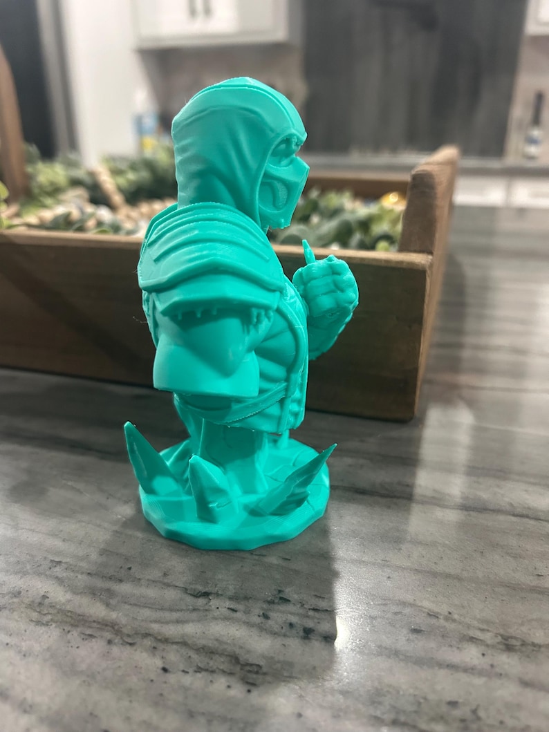 May include: A bright teal 3D printed bust of a hooded warrior figure wearing a mask and shoulder armour. The figure is depicted from the chest up, posed with a clenched fist, and stands on a base featuring jagged, crystalline-like protrusions.