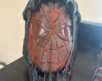 May include: A three-dimensional sculpted head featuring a red and black textured mask with a web-like pattern, encased within a dark black, jagged, toothy outer shell that frames the face with sharp, pointed protrusions at the top and bottom.