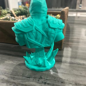 May include: A bright teal 3D printed bust of a figure wearing armour with shoulder plates and straps. The figure is depicted from the back, showing a hooded head and a textured torso. The base of the bust features jagged, crystal-like protrusions.