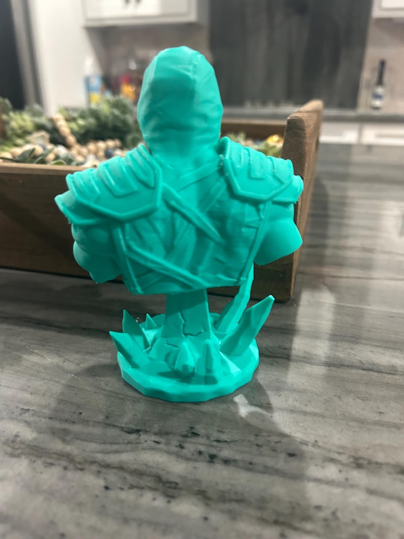 May include: A bright teal 3D printed bust of a figure wearing armour with shoulder plates and straps. The figure is depicted from the back, showing a hooded head and a textured torso. The base of the bust features jagged, crystal-like protrusions.