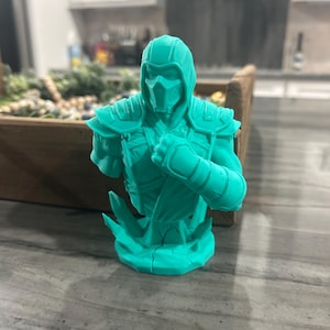 May include: A monochromatic teal 3D printed bust of a masked warrior figure wearing a hooded garment and shoulder armour. The figure is posed with one arm raised in a defensive stance, set upon a jagged, crystalline-textured base structure.