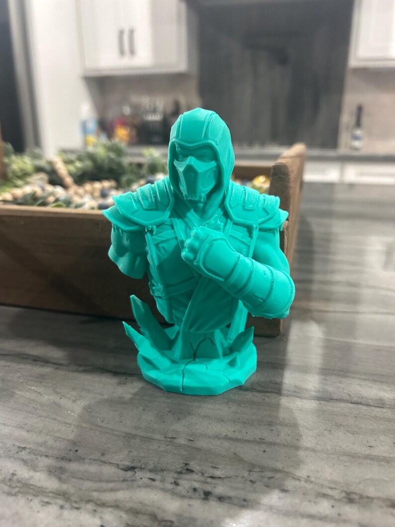 May include: A monochromatic teal 3D printed bust of a masked warrior figure wearing a hooded garment and shoulder armour. The figure is posed with one arm raised in a defensive stance, set upon a jagged, crystalline-textured base structure.