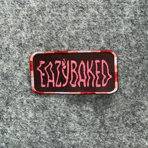 May include: A rectangular embroidered patch with a dark brown background and a red and white checkered border. The text EAZYBAKED is stitched in a stylized, wavy pink font across the center of the patch, which features rounded corners.