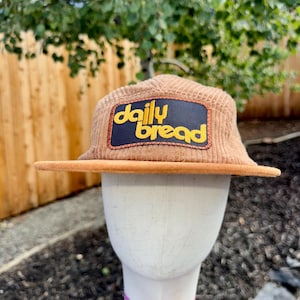 Op de afbeelding: Een beige corduroy baseballpet met een rechthoekige patch op het voorpaneel. De donkerblauwe patch met een oranje gestikte rand bevat de gele tekst daily bread in een afgerond, kleine lettertype. De pet heeft een platte, beige klep.