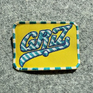 May include: A rectangular embroidered patch with a bright yellow background and a white and teal striped border. The text GRIZ is stitched in the center in a stylized font with white and teal diagonal stripes and a dark blue outline.