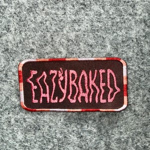 May include: A rectangular embroidered patch with a dark brown background and a pink stitched text that reads EAZYBAKED. The patch features a border with a repeating pattern of red, orange, and white vertical stripes along the edges.