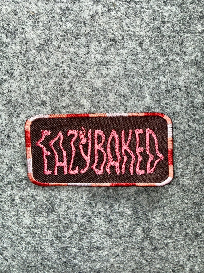 May include: A rectangular embroidered patch with a dark brown background and a pink stitched text that reads EAZYBAKED. The patch features a border with a repeating pattern of red, orange, and white vertical stripes along the edges.