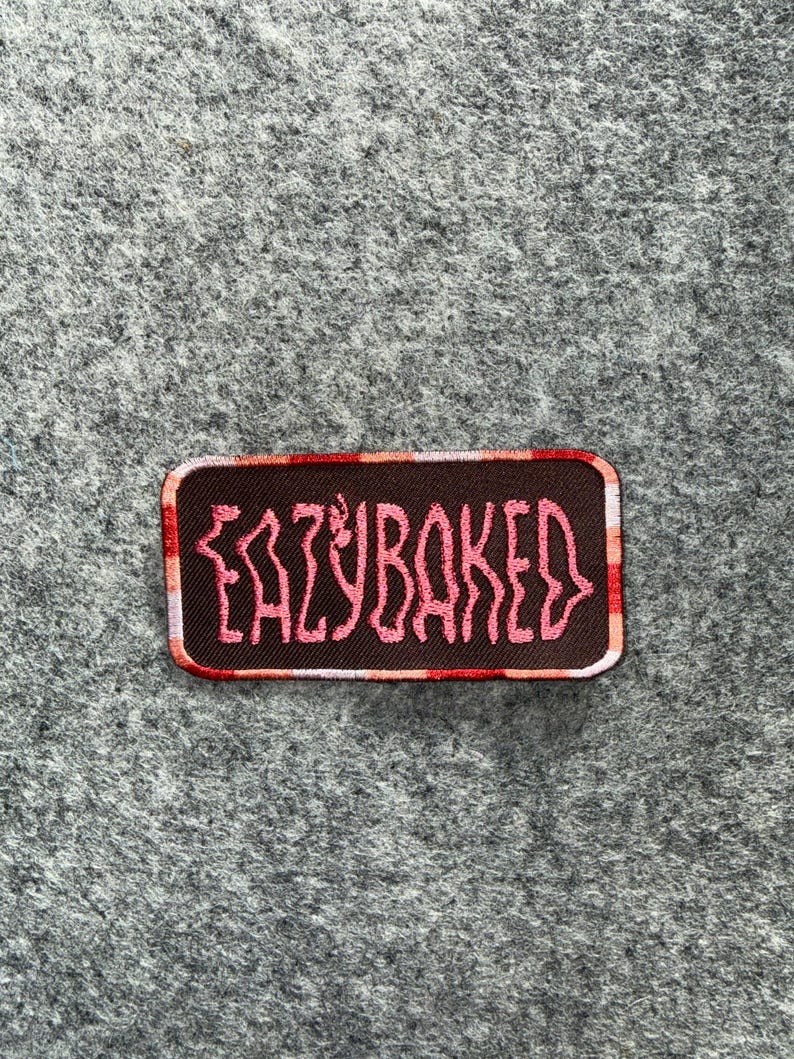May include: A rectangular embroidered patch with a dark brown background and a red and white checkered border. The text EAZYBAKED is stitched in a stylized, wavy pink font across the center of the patch, which features rounded corners.