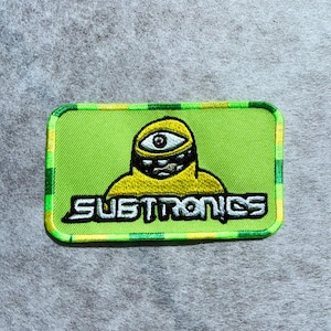 May include: A rectangular embroidered patch with a lime green background and a checkered border in yellow and green. The patch features a yellow cyclops character with a single eye and a wide mouth, positioned above the white text SUBTRONICS.