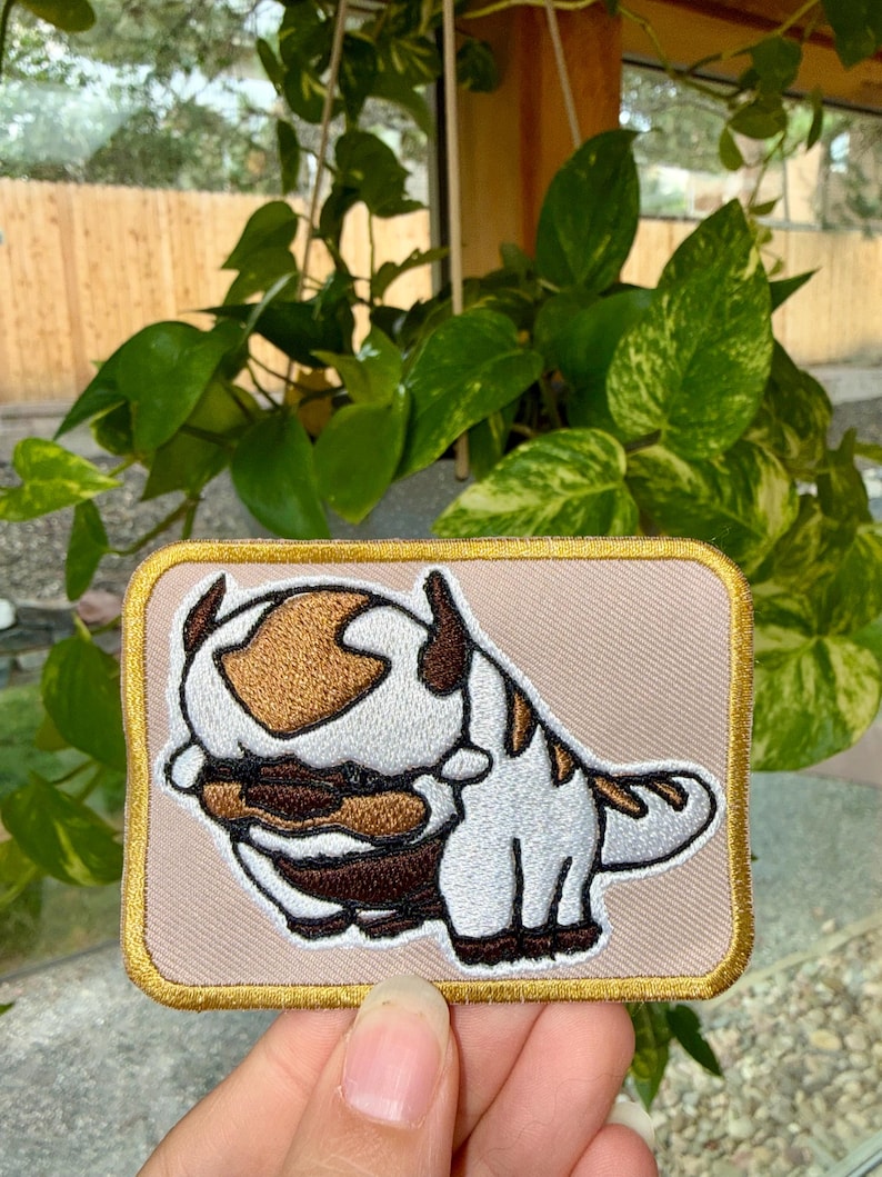 May include: A rectangular embroidered patch featuring a white and brown creature with a large head, pointed ears, and a brown arrow marking on its forehead. The patch has a light beige background and a thick, metallic gold stitched border.