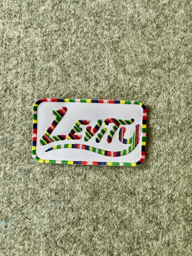 Peut inclure: Un &eacute;cusson rectangulaire brod&eacute; avec un fond blanc et une bordure ray&eacute;e multicolore. Le mot Levity est cousu au centre avec un motif de fil ray&eacute; multicolore assorti &agrave; la bordure, pr&eacute;sentant un style d'&eacute;criture cursive manuscrite.