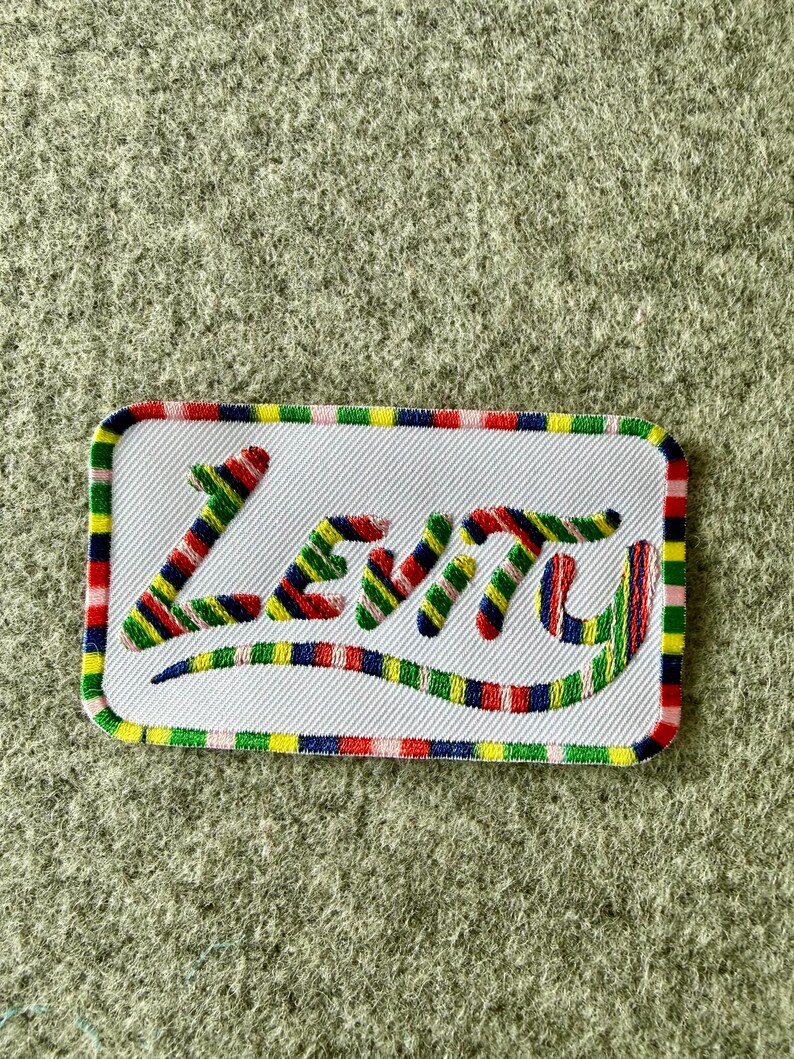Peut inclure: Un &eacute;cusson rectangulaire brod&eacute; avec un fond blanc et des coins arrondis. Le mot Levity est cousu dans un motif ray&eacute; multicolore compos&eacute; de rouge, bleu, jaune et vert. La bordure pr&eacute;sente un motif r&eacute;p&eacute;titif de ces m&ecirc;mes couleurs vives.