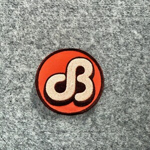 May include: A circular embroidered patch featuring a vibrant orange background with a dark brown border. In the centre, a stylised, cream-coloured letter B is outlined in dark brown, creating a bold, graphic design with a raised, textured appearance.