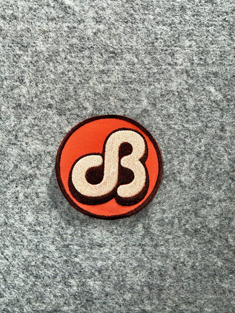 May include: A circular embroidered patch featuring a vibrant orange background with a dark brown border. In the centre, a stylised, cream-coloured letter B is outlined in dark brown, creating a bold, graphic design with a raised, textured appearance.