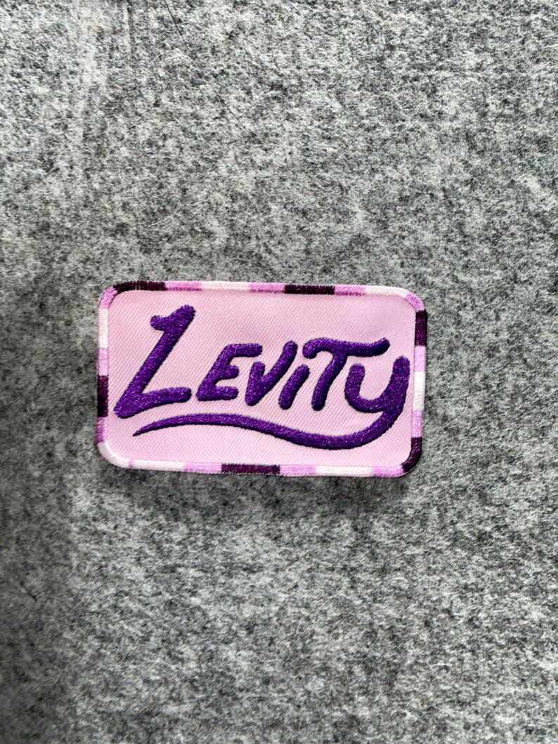 Peut inclure: Un &eacute;cusson rectangulaire brod&eacute; pr&eacute;sentant un fond rose clair avec une bordure &agrave; carreaux violet fonc&eacute;. Le mot Levity est brod&eacute; dans une police cursive grasse de couleur violet fonc&eacute; au centre de l'&eacute;cusson, cr&eacute;ant un contraste visuel net.
