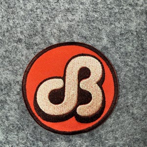 May include: A circular embroidered patch featuring a bright orange background with a dark brown border. In the centre, a stylised, cream-coloured letter B is outlined in dark brown, creating a bold, graphic design with a raised, textured appearance.
