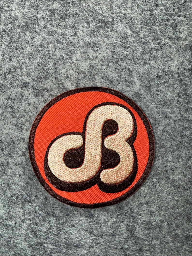 May include: A circular embroidered patch featuring a bright orange background with a dark brown border. In the centre, a stylised, cream-coloured letter B is outlined in dark brown, creating a bold, graphic design with a raised, textured appearance.