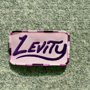 Peut inclure: Un &eacute;cusson rectangulaire brod&eacute; pr&eacute;sentant un fond rose clair avec une bordure &agrave; carreaux violets et noirs. Le mot Levity est cousu en fil violet fonc&eacute; au centre dans une police stylis&eacute;e inspir&eacute;e de l'&eacute;criture cursive avec une fioriture.