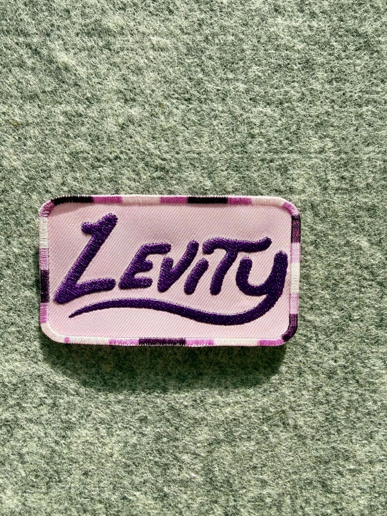 Peut inclure: Un &eacute;cusson rectangulaire brod&eacute; pr&eacute;sentant un fond rose clair avec une bordure &agrave; carreaux violets et noirs. Le mot Levity est cousu en fil violet fonc&eacute; au centre dans une police stylis&eacute;e inspir&eacute;e de l'&eacute;criture cursive avec une fioriture.