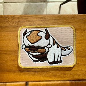 Puede incluir: Un parche bordado rectangular que presenta una criatura estilizada de color blanco y marr&oacute;n con una gran marca en forma de flecha en la cabeza. El parche tiene un fondo beige claro y un borde dorado, mostrando costuras detalladas en el dise&ntilde;o.