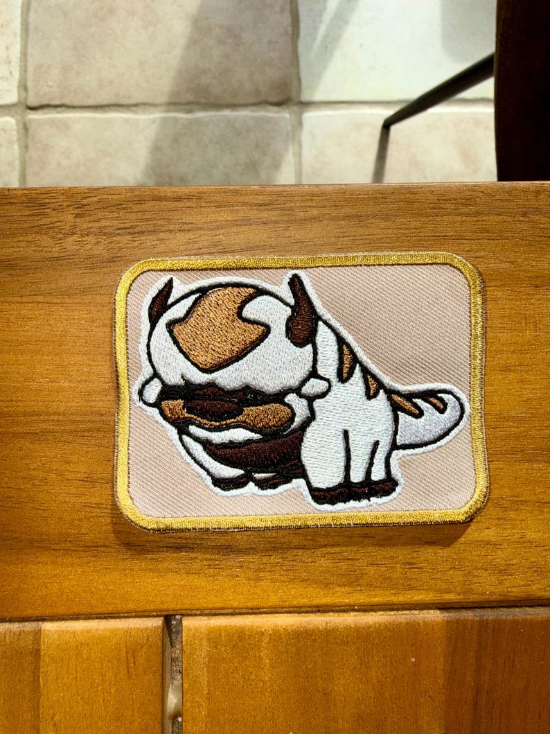Puede incluir: Un parche bordado rectangular que presenta una criatura estilizada de color blanco y marr&oacute;n con una gran marca en forma de flecha en la cabeza. El parche tiene un fondo beige claro y un borde dorado, mostrando costuras detalladas en el dise&ntilde;o.