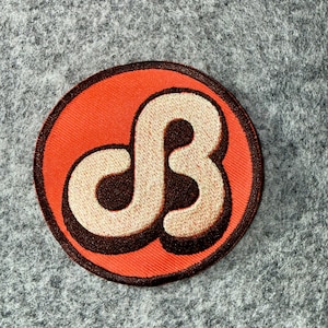May include: A circular embroidered patch featuring a bright orange background with a dark brown border. In the centre, a stylised, cream-coloured letter B is depicted with a dark brown drop shadow, creating a bold and graphic appearance.