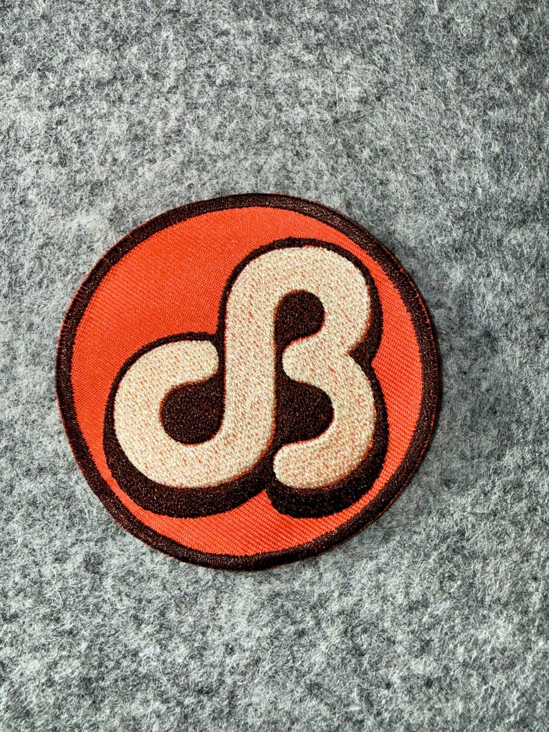May include: A circular embroidered patch featuring a bright orange background with a dark brown border. In the centre, a stylised, cream-coloured letter B is depicted with a dark brown drop shadow, creating a bold and graphic appearance.