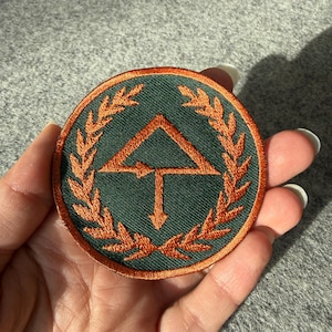 May include: A circular embroidered patch featuring a dark green background with a copper-colored border. The design includes a central geometric arrow symbol pointing upward and downward, surrounded by a detailed copper-colored laurel wreath.