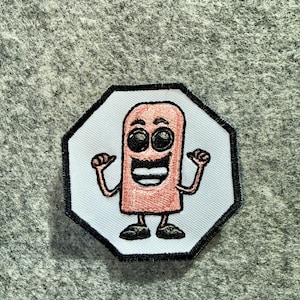 Peut inclure: Un écusson brodé octogonal représentant un personnage de dessin animé au corps rose rectangulaire, avec de grands yeux noirs, une large bouche souriante et des bras fins pointant vers sa poitrine, le tout sur fond blanc.