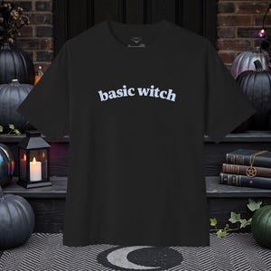 May include: A black short-sleeved t-shirt featuring the text "basic witch" printed in white across the chest. The shirt has a crew neckline and a standard fit, constructed from a solid black fabric that provides a neutral base for the lettering.