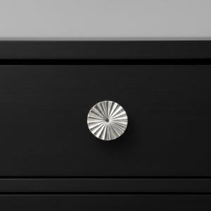 Puede incluir: Un pomo de mueble circular de metal plateado con un patrón de rayos de sol formado por crestas radiantes. El pomo está montado en el centro de un frente de cajón negro mate, que presenta líneas horizontales hundidas arriba y abajo.