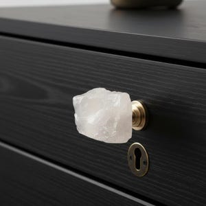 May include: A drawer knob featuring a raw, translucent white quartz stone attached to a circular brass-toned metal base. The knob is mounted on a black wooden drawer front that also includes a matching brass-toned metal keyhole escutcheon plate.