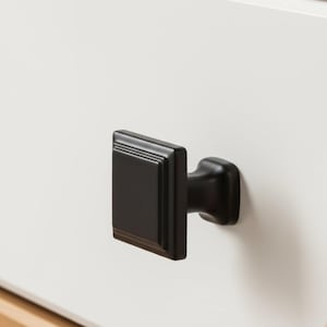 May include: A matte black cabinet knob with a square face and stepped edges, mounted on a plain white surface. The hardware features a smooth, solid finish with a central rectangular panel and a sturdy, tapered base connecting to the drawer front.