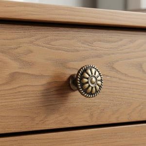 May include: A light brown wooden drawer front featuring a prominent wood grain pattern. Attached to the center is a round, antique brass-toned metal knob with a raised floral petal design and a decorative rope-style border around the edge.
