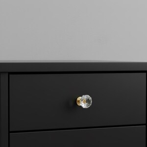 May include: A matte black drawer front featuring a single faceted clear glass knob with a gold-toned metal base. The knob is centered on the drawer, which is part of a larger piece of furniture with a smooth, dark finish and clean lines.