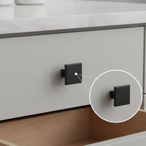 May include: A matte black square cabinet knob with a flat front surface and a central post, shown attached to a light gray drawer front, with an additional circular inset highlighting the knob from a slightly different side angle.