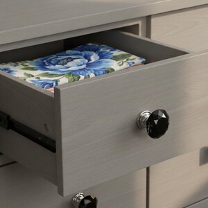 May include: A light gray wooden dresser with an open drawer containing a folded fabric featuring a blue and white floral pattern. The drawer front is fitted with a faceted black crystal-style knob with a silver base, matching the lower drawer knob.