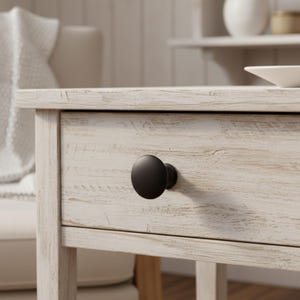 May include: A matte black circular cabinet knob is mounted on the front of a light-colored wooden drawer. The drawer features a distressed, weathered finish with visible wood grain patterns and a horizontal orientation on a piece of furniture.