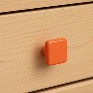 May include: A square, bright orange cabinet knob with rounded corners attached to a light-colored wooden drawer front. The wood grain is visible, showing horizontal lines across the surface of the drawer, while the knob has a smooth finish.