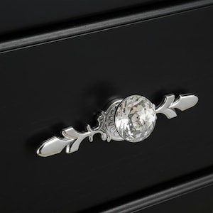 May include: A drawer pull handle featuring a central clear, faceted, round crystal knob mounted on a polished silver-tone metal base. The metal base has an ornate, elongated design with pointed ends, attached to a solid matte black drawer front.