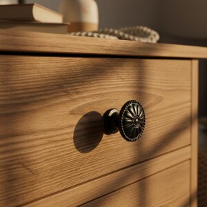 Op de afbeelding: Een ronde, donkergekleurde metalen ladeknop met een decoratief, radiaal bloembladpatroon en een kralenrand, gemonteerd op een lichtgekleurd houten ladefront met een zichtbare houtnerf en horizontale paneeldetails.