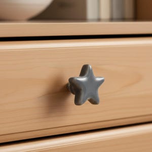 May include: A star-shaped drawer knob with a smooth, glossy grey finish. The knob is attached to a light-toned wooden drawer front, featuring a simple five-pointed design with rounded edges and a central mounting point connecting to the wood.