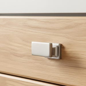 May include: A rectangular, brushed silver metal cabinet knob with a smooth, flat front surface and a stepped base, mounted onto a light-coloured, horizontal wood grain drawer front with a visible natural texture and light brown wood tones.