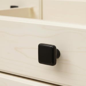 May include: A square, glossy black cabinet knob attached to a light-coloured wooden drawer front. The knob features rounded corners and a smooth, reflective surface, contrasting with the natural grain pattern of the pale wood drawer panel.